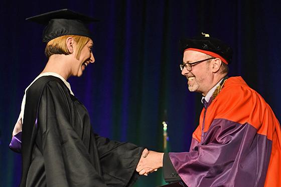 图为菲尼戈尔德校长在台上与一名微笑的学生握手, 都穿着毕业袍，戴着毕业帽