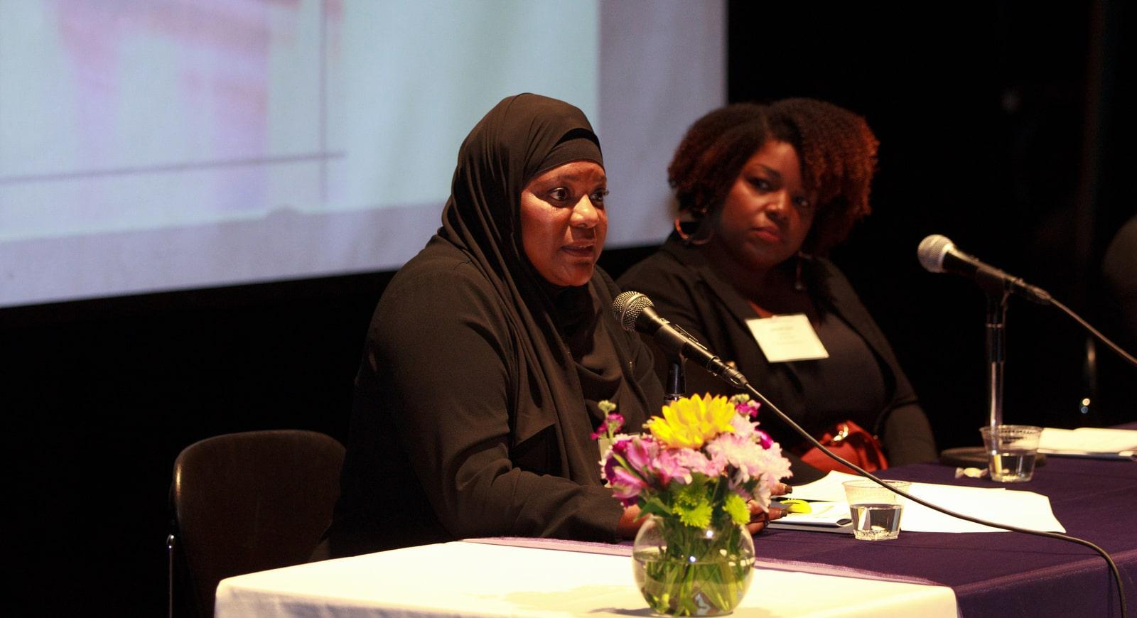 Photo of two Black women on a panel. The hijabi woman is speaking, while the woman beside her listens.